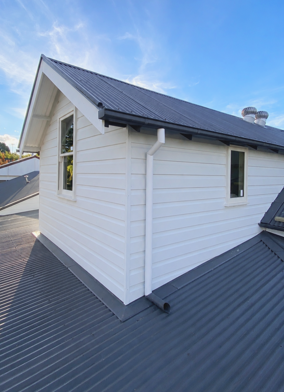 A white house with a black metal roof, two small windows, and a white gutter system. The sky is blue with some clouds.
