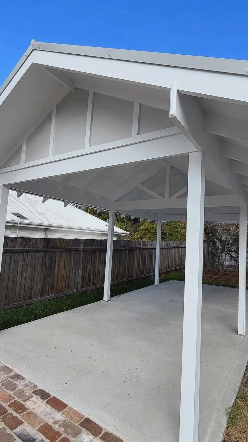 A white modern outdoor patio cover with a gabled roof, supported by white posts, over a concrete slab in a backyard, with a wooden fence and neighboring house in the background.
