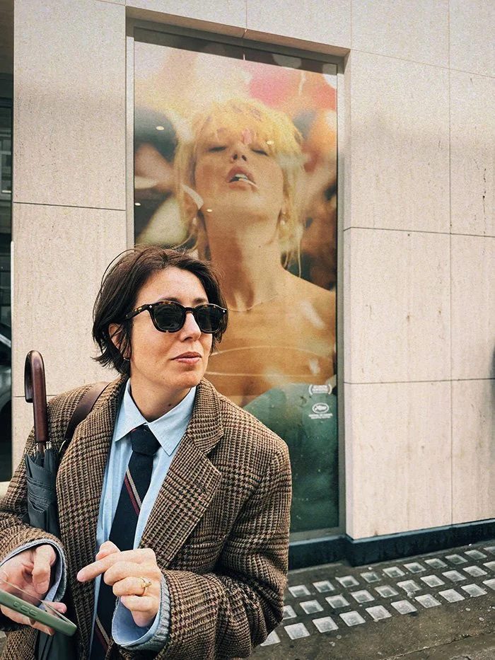 A woman with short dark hair, wearing sunglasses, a checkered blazer, and a tie, holds a phone and an umbrella as she stands outside a building with a large advertisement featuring a woman with blond hair and closed eyes behind her.