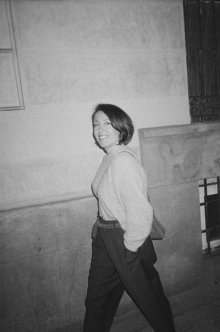A woman with short hair smiling and walking outdoors at night, wearing a hoodie and high-waisted pants, with her hands in her pockets.