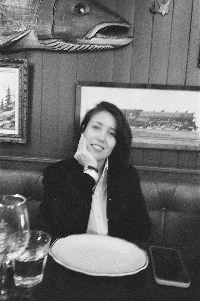 A woman sitting at a table in a restaurant, smiling, with a fish head mounted on the wall behind her, along with framed pictures and wood-paneled walls.