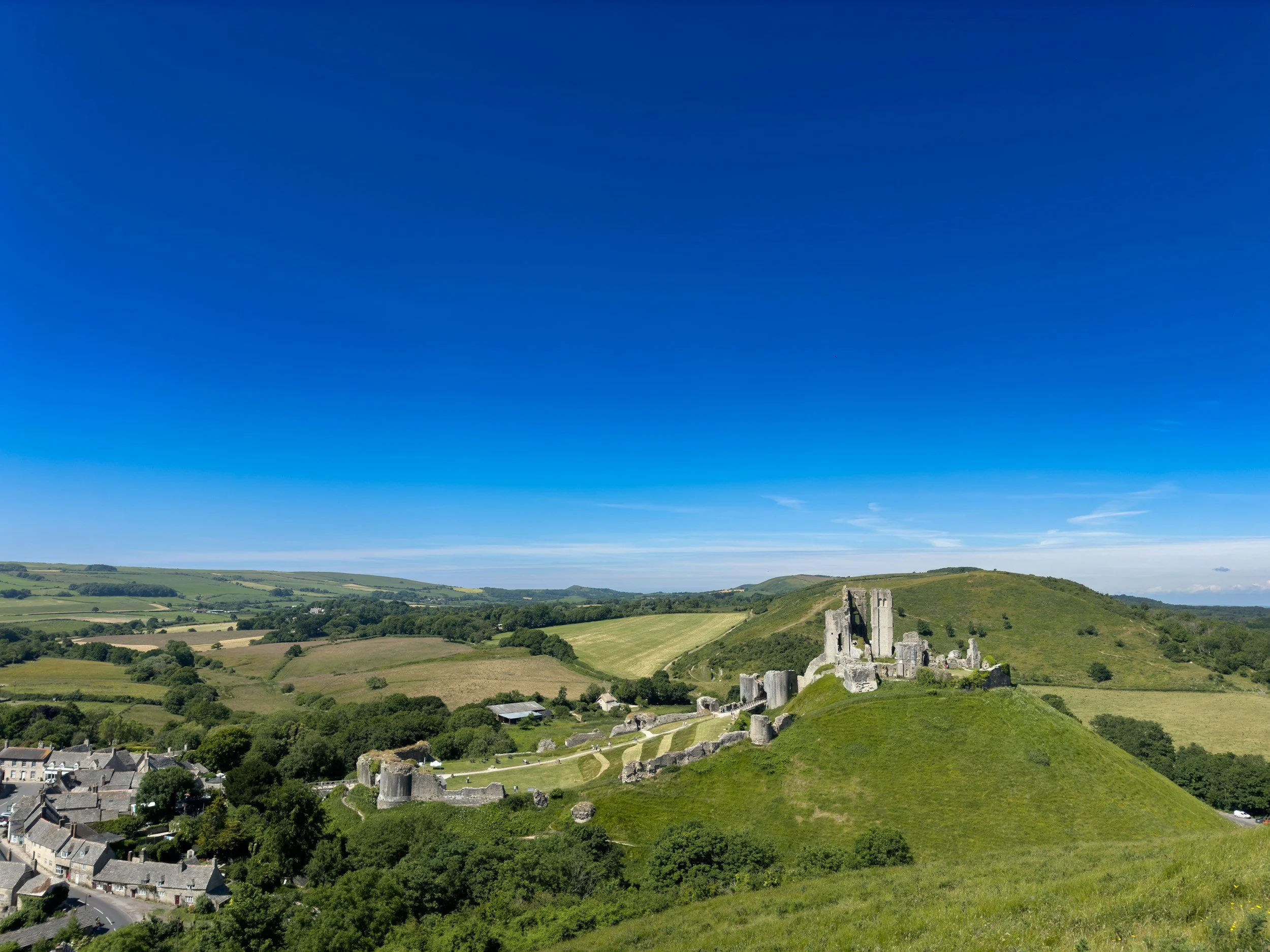 A landscape view of rolling green hills with ruins of an ancient castle and stone fortress walls, under a clear blue sky.