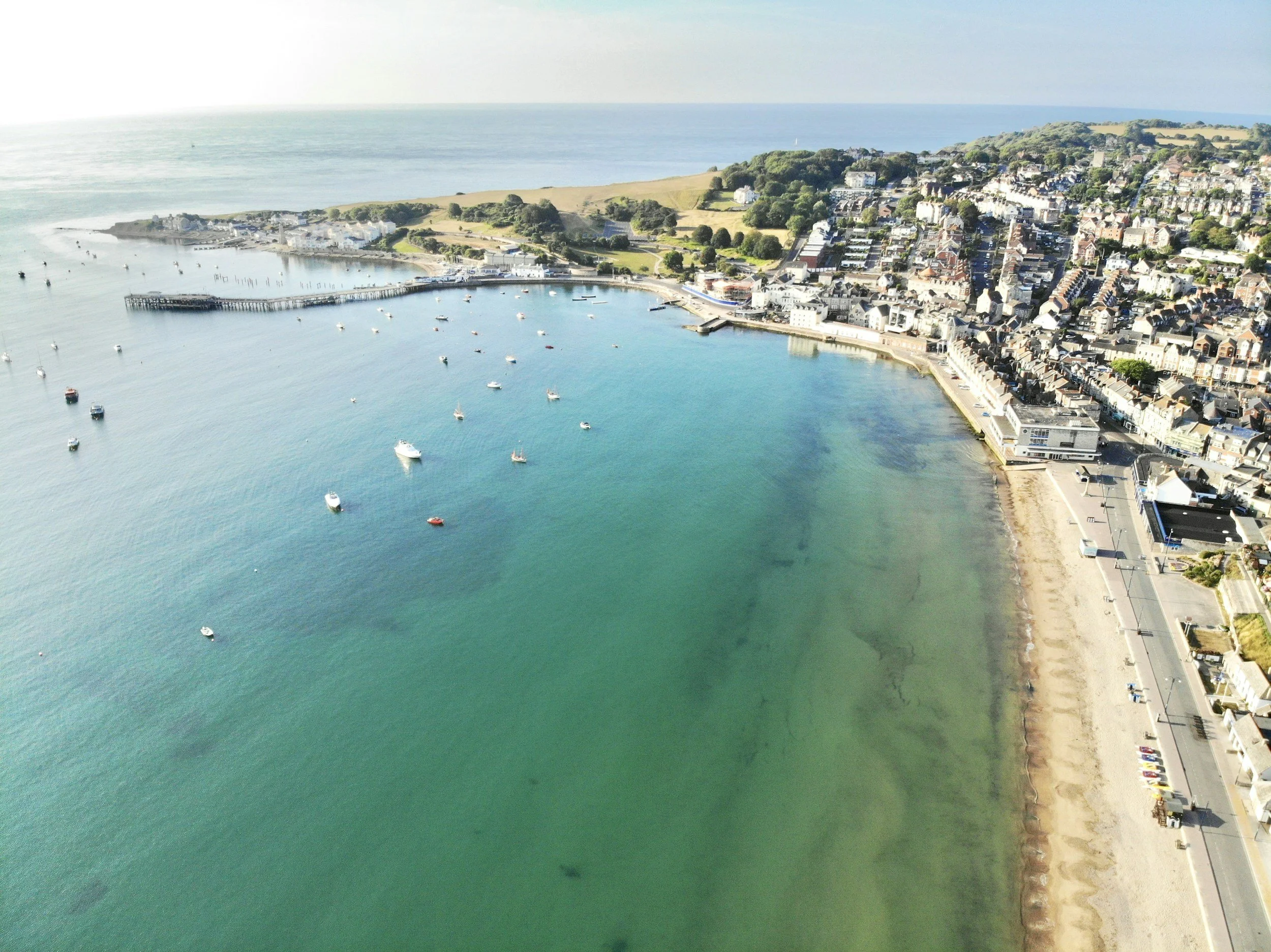 An aerial view of a coastal town with a harbor filled with boats, a sandy beach, and densely packed buildings. The ocean and green hills are in the background.