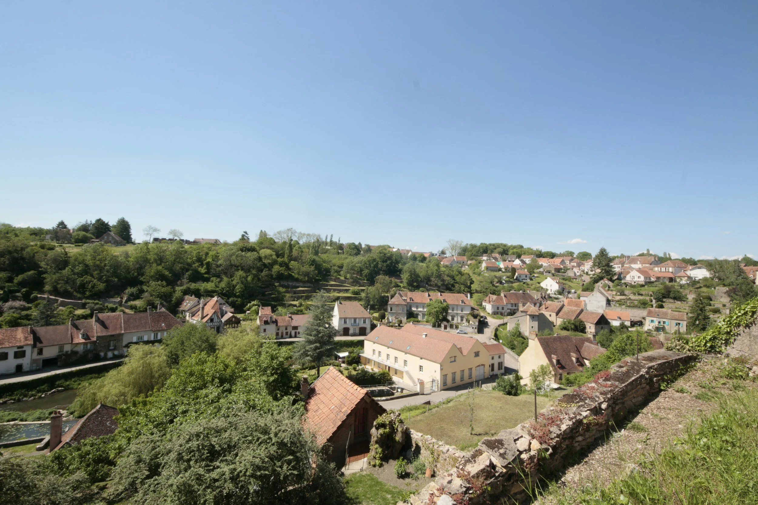 A scenic view of a small town or village with houses, surrounded by lush green trees and hills under a clear blue sky.