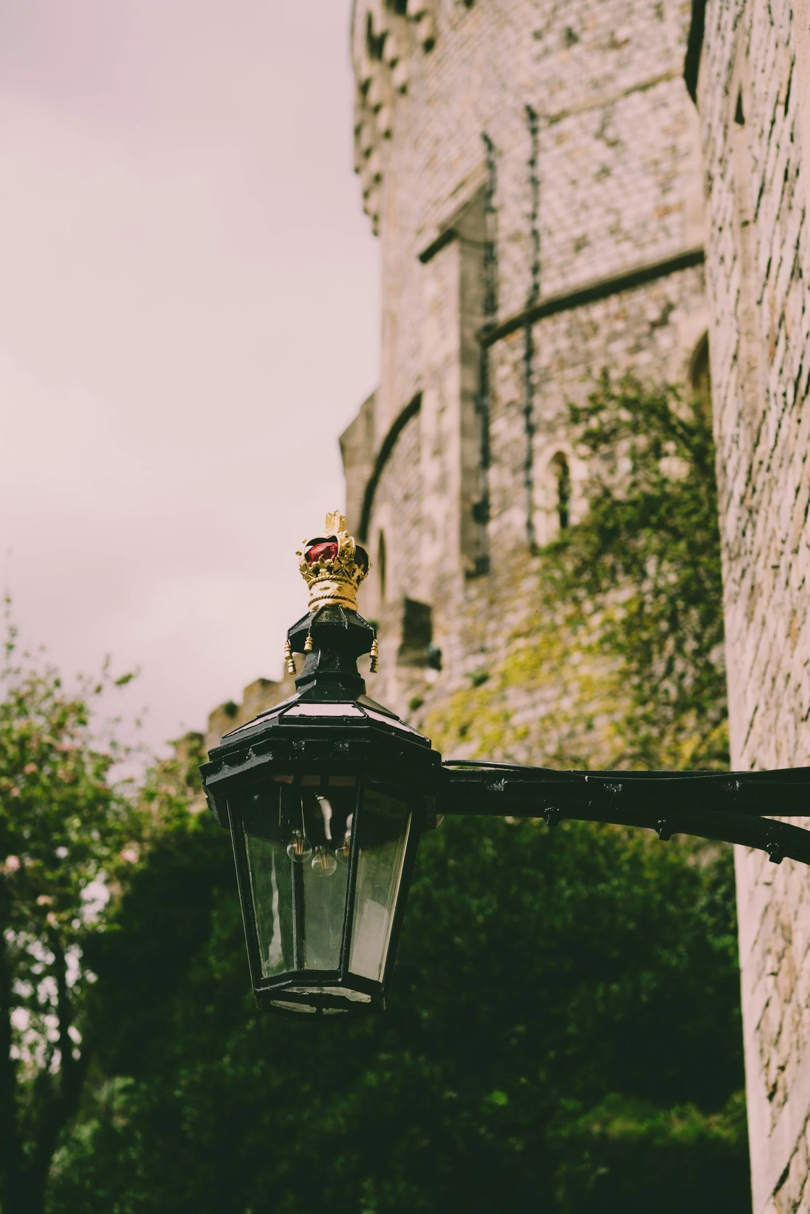 A black street lamp with golden accents and a crown motif on top, hanging in front of an old stone building with trees and a cloudy sky in the background.