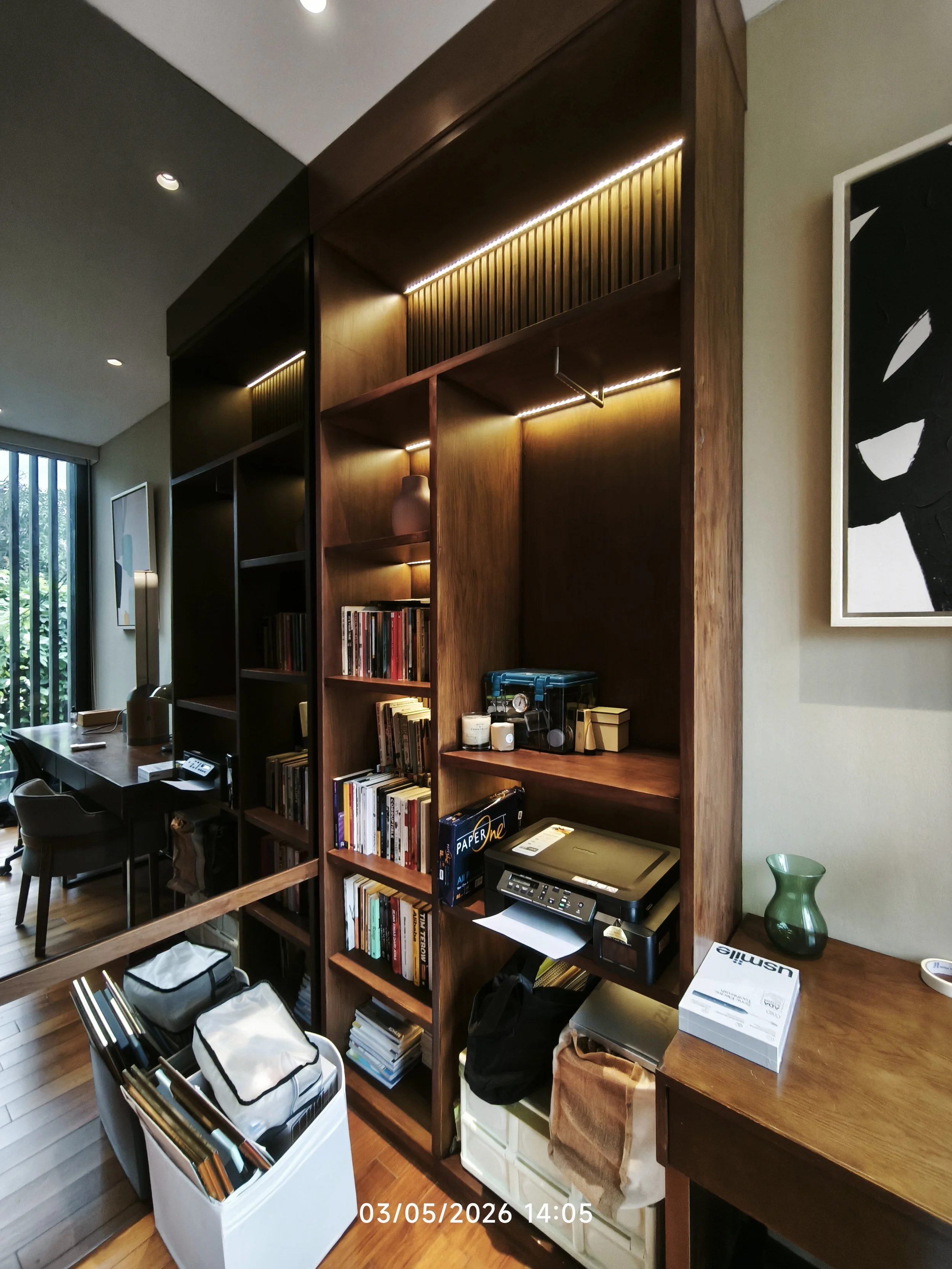 A room with wooden shelves filled with books and decorative items, a desk with office supplies, a window with blinds letting in natural light, and various storage bins and boxes.
