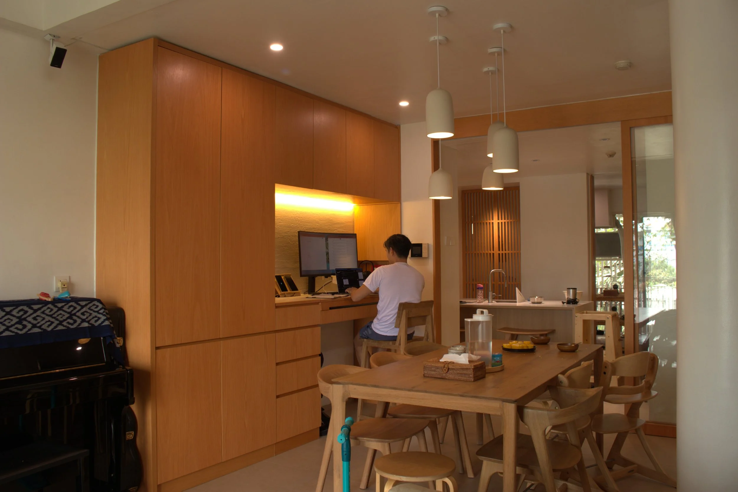 A person sitting at a desk with a computer in a modern kitchen or dining area with wooden cabinetry and pendant lights.