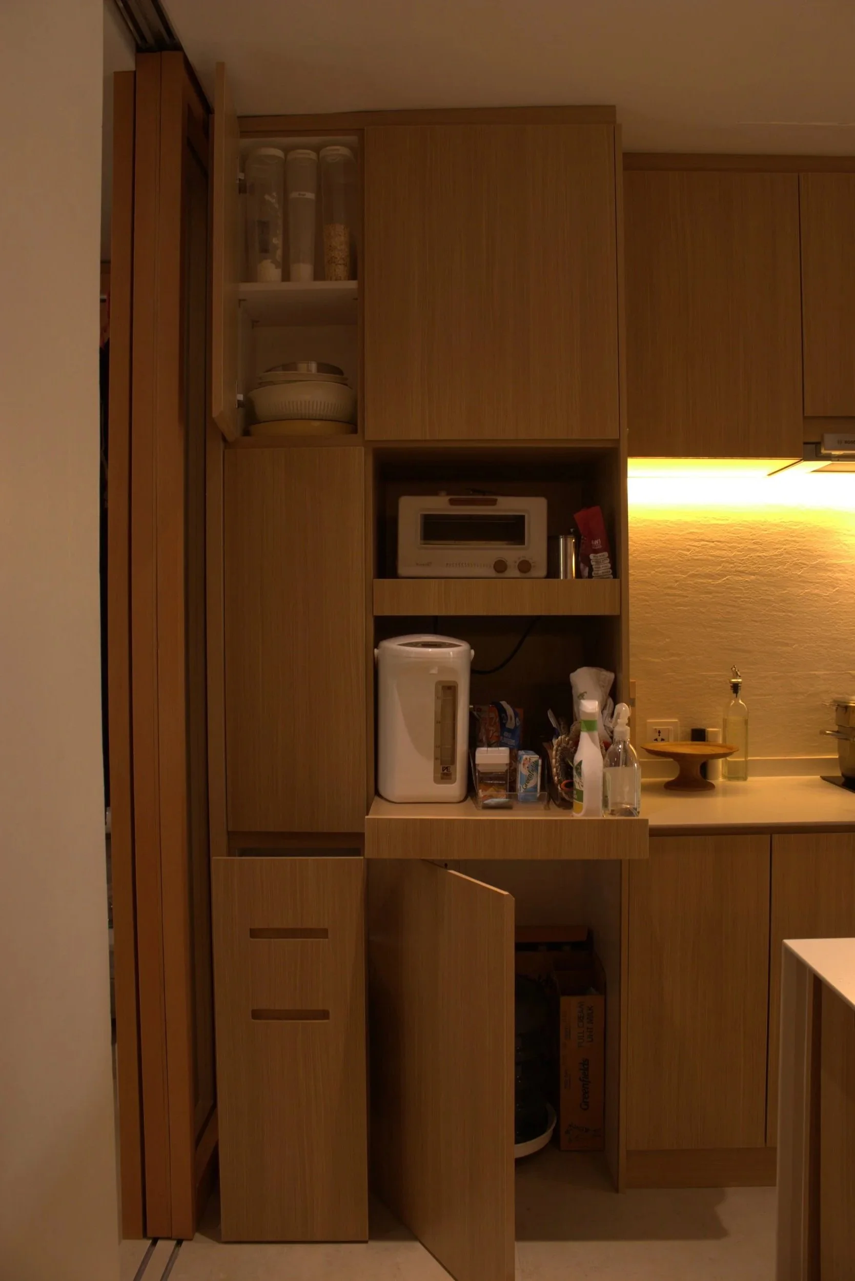 Kitchen cabinets with various appliances and utensils, including a microwave, toaster, and water filter, illuminated by warm yellow lighting.