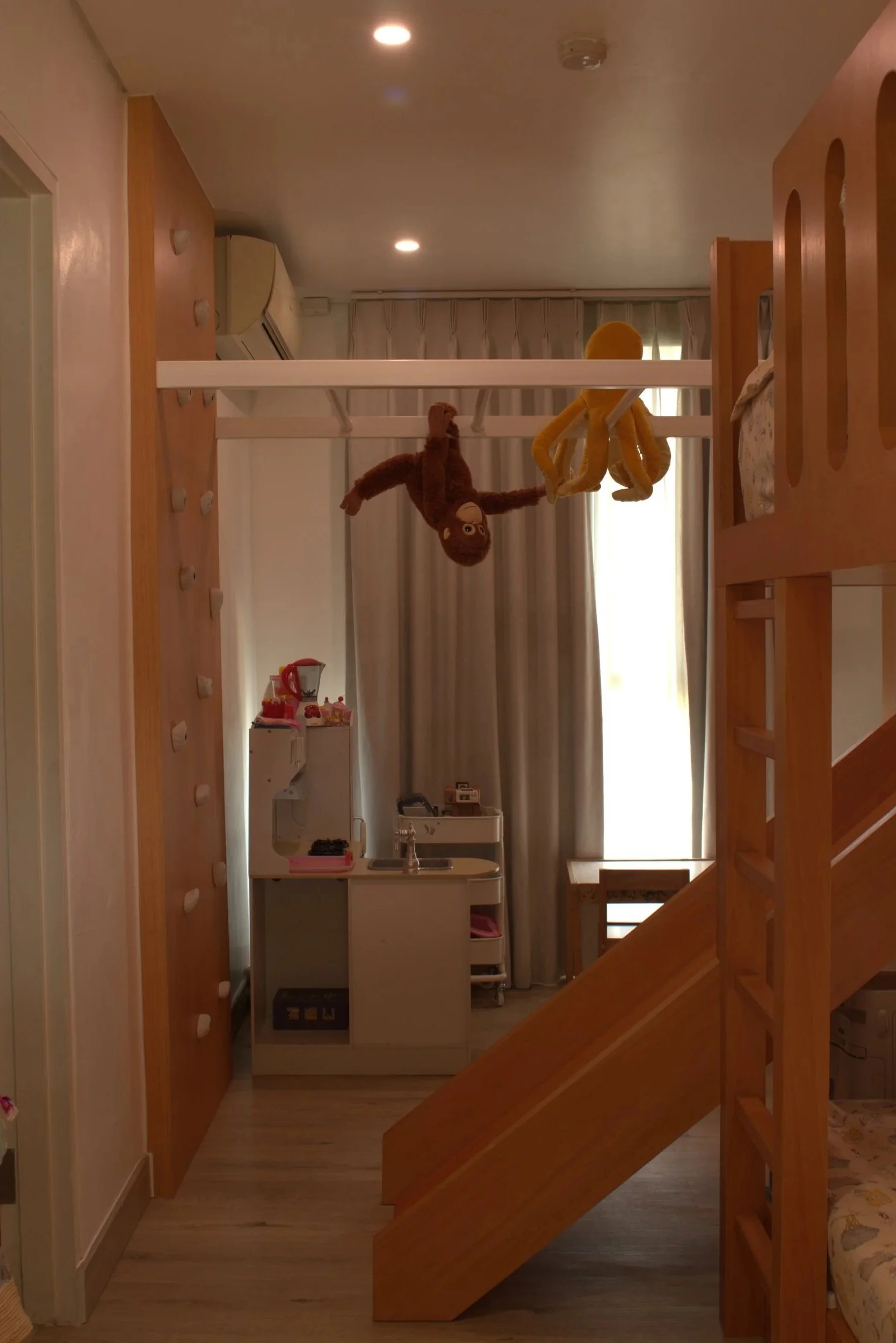 A small child's play area with a loft bed, hanging plush monkey and octopus toys, a miniature kitchen set underneath, and wooden stairs. Curtains cover a window, and there are ceiling lights.