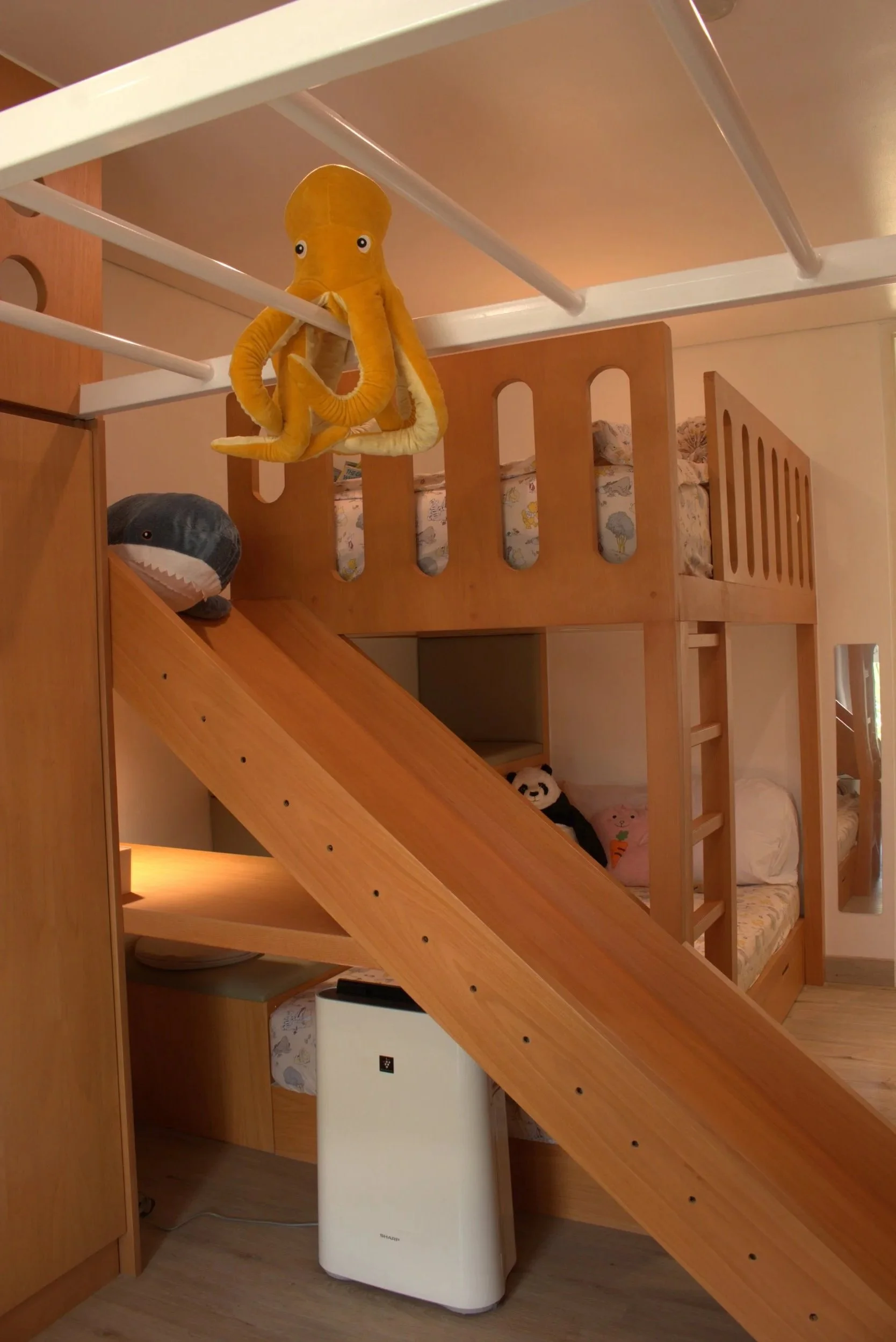 The image shows a loft bed with a wooden ladder. A yellow plush octopus toy is hanging from a white clothing rack or shelf above. There is a gray plush shark toy on a bed or couch underneath, and a white air purifier or dehumidifier is positioned bel