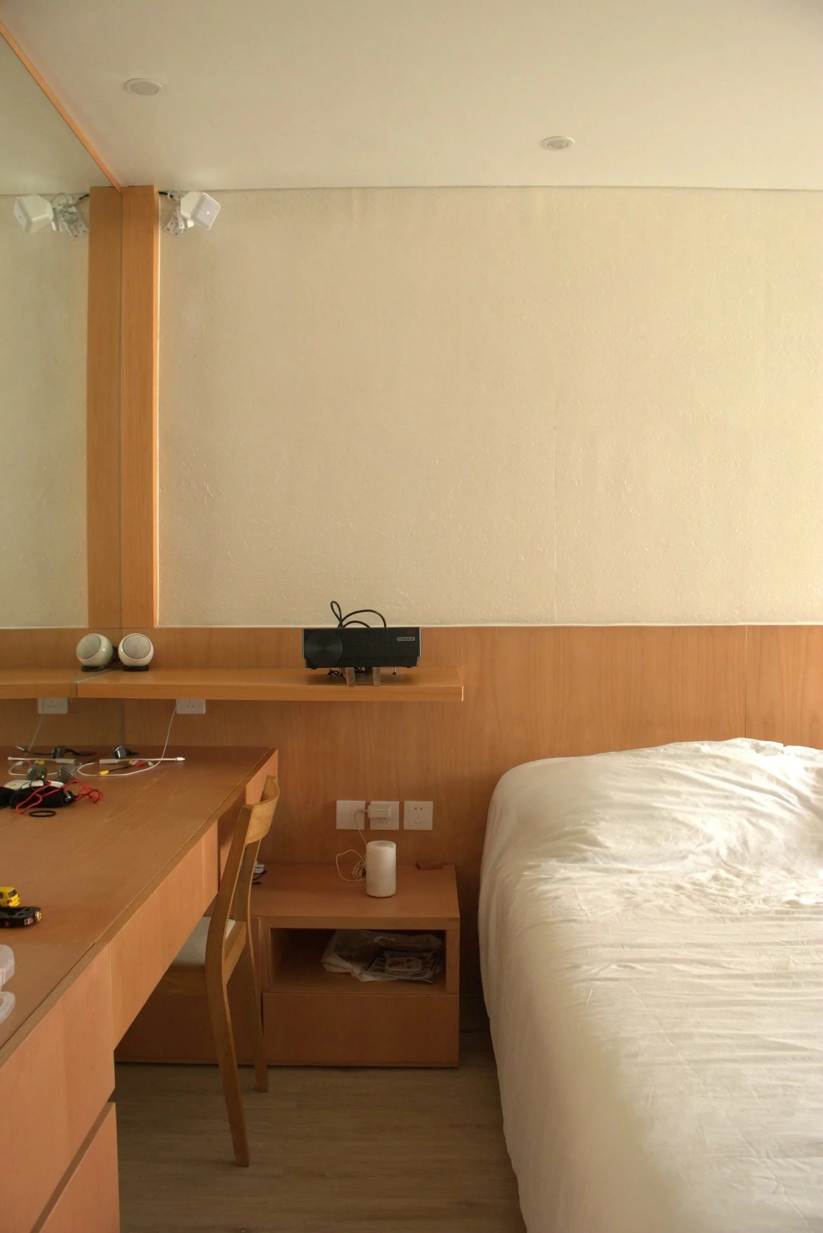 A hotel room with a bed, wooden furniture, a wall-mounted mirror, a small desk, and electronic devices including a speaker and a remote, with a textured beige wall and wood flooring.