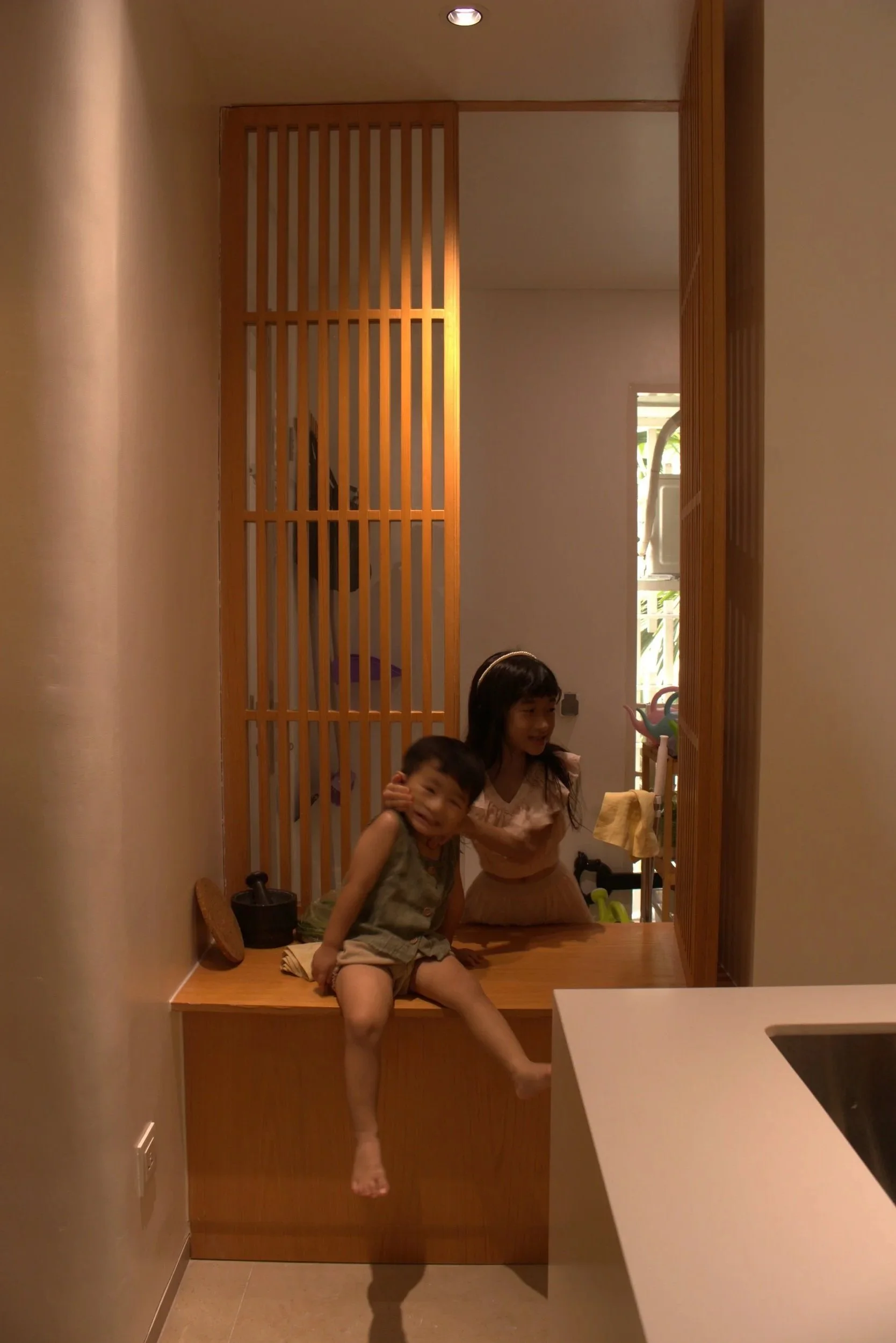 Two children, a boy and a girl, sitting on a wooden bench in a room with wooden sliding doors, with the boy hugging the girl and both smiling.