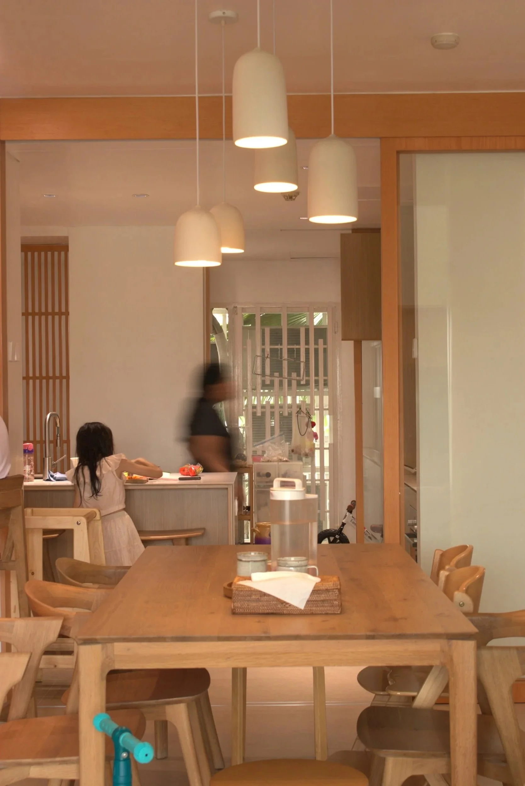 Interior of a modern, minimalist restaurant or cafe with wooden furniture, hanging pendant lamps, and a large dining table in the foreground. A child is sitting at the table, and a blurred person is walking in the background near a window.