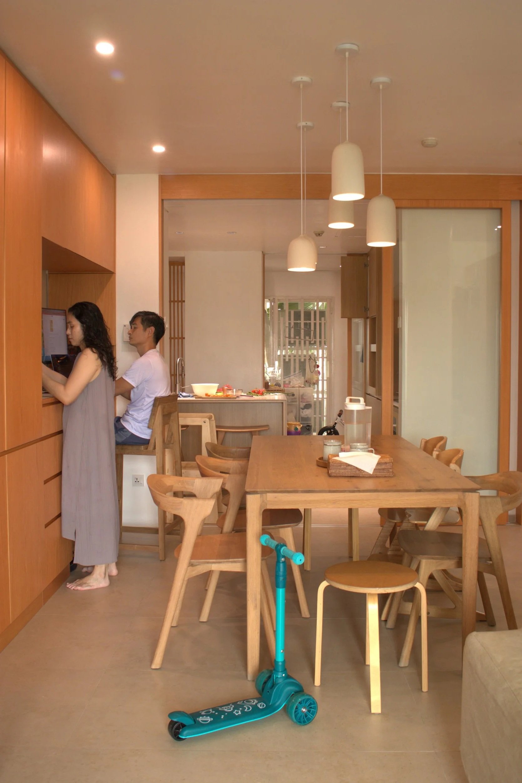 A kitchen with wooden furniture, a long dining table with chairs, a woman and a man at the counter, and a blue children's scooter on the floor.