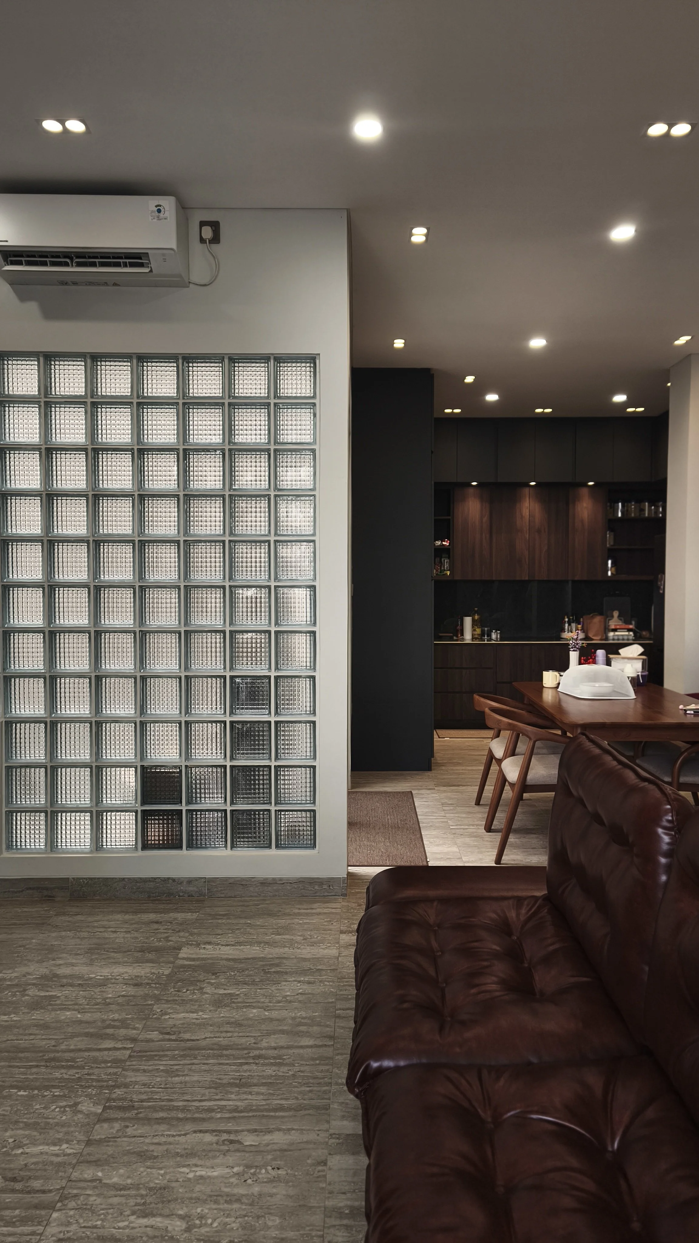A living room with brown leather sofa, glass block wall on the left, wooden dining table in the background, and black kitchen cabinets. Ceiling with multiple recessed lights, air conditioning unit on the left.