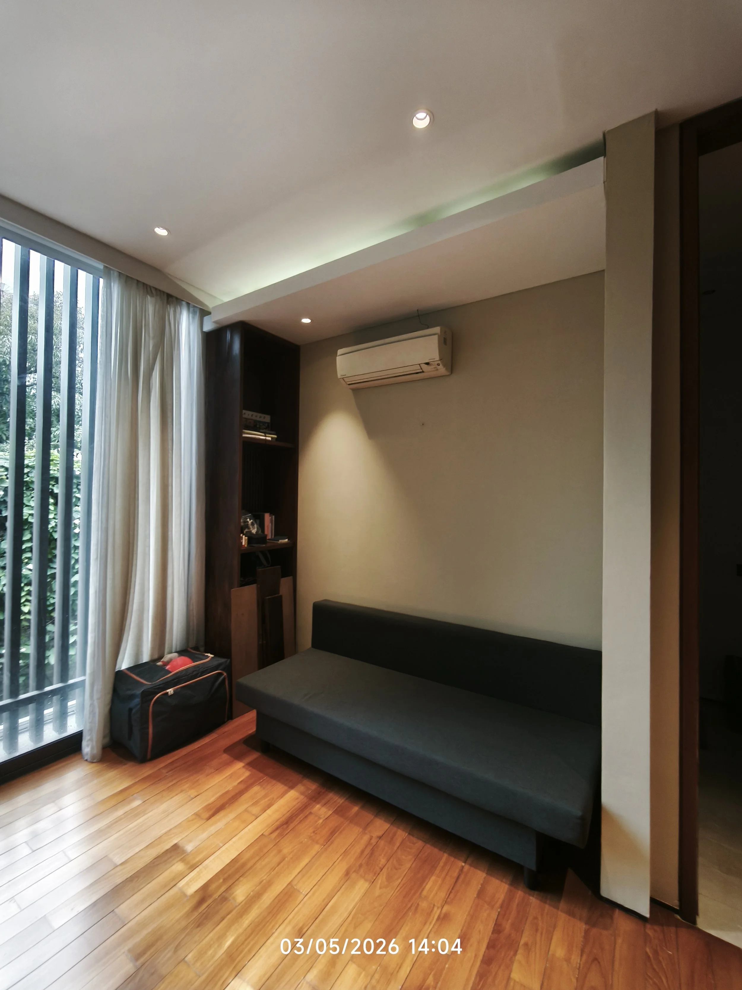 A living room corner with a sliding glass door, white curtains, wooden flooring, a black sofa, a small black suitcase, a dark wooden bookcase with books, and a wall-mounted air conditioning unit.
