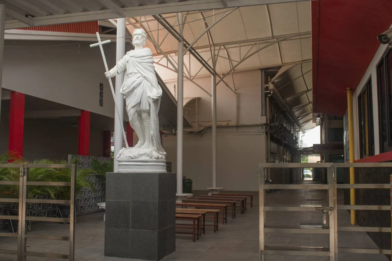 White statue of Jesus Christ holding a cross, situated on a black pedestal, inside a covered outdoor area with benches and modern architectural features.