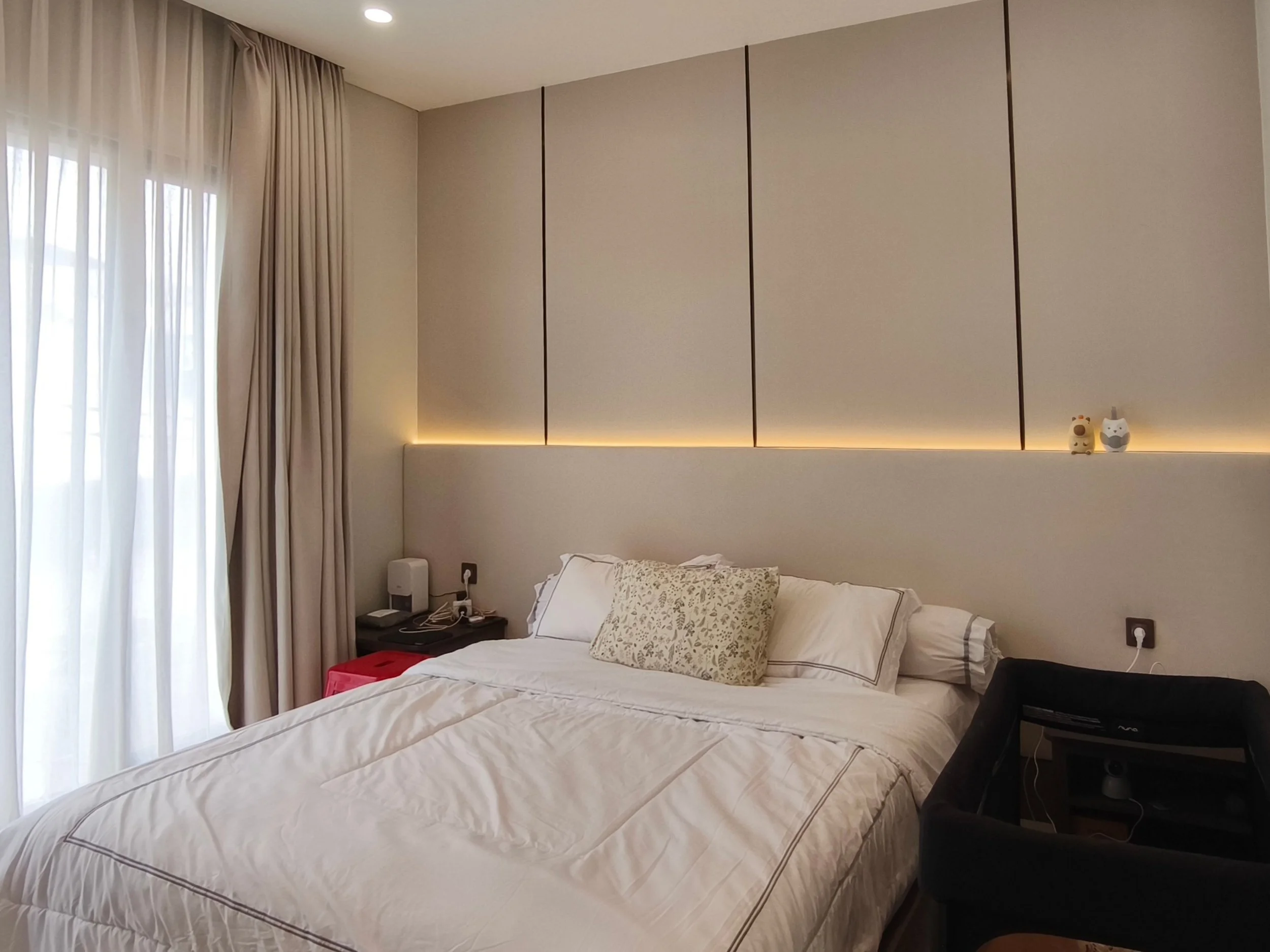 A bedroom with two neatly made beds, beige curtains on the window, and a beige decorative wall panel with LED lighting. Two small decorative figurines sit on a ledge above the bed, and there are bedside tables with electronic devices.