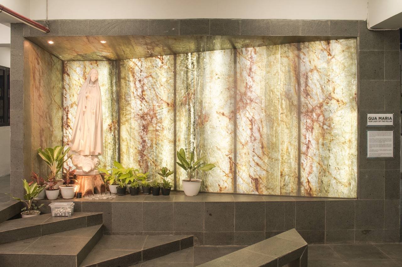 Statue of the Virgin Mary on a pedestal with potted plants in front, set against a backlit onyx wall in a religious display area.