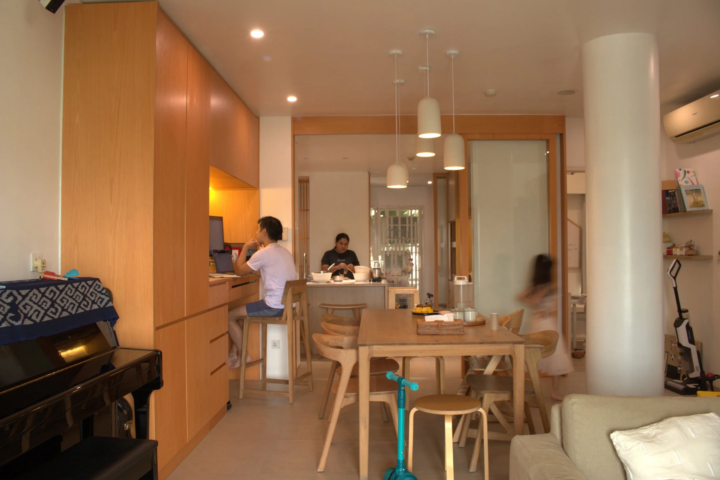 A cozy, modern kitchen and dining area with warm wooden accents. There are two people seated at the counter, one working on a laptop and the other preparing food. The space has pendant lighting, a dining table with several chairs, and a piano in the corner.