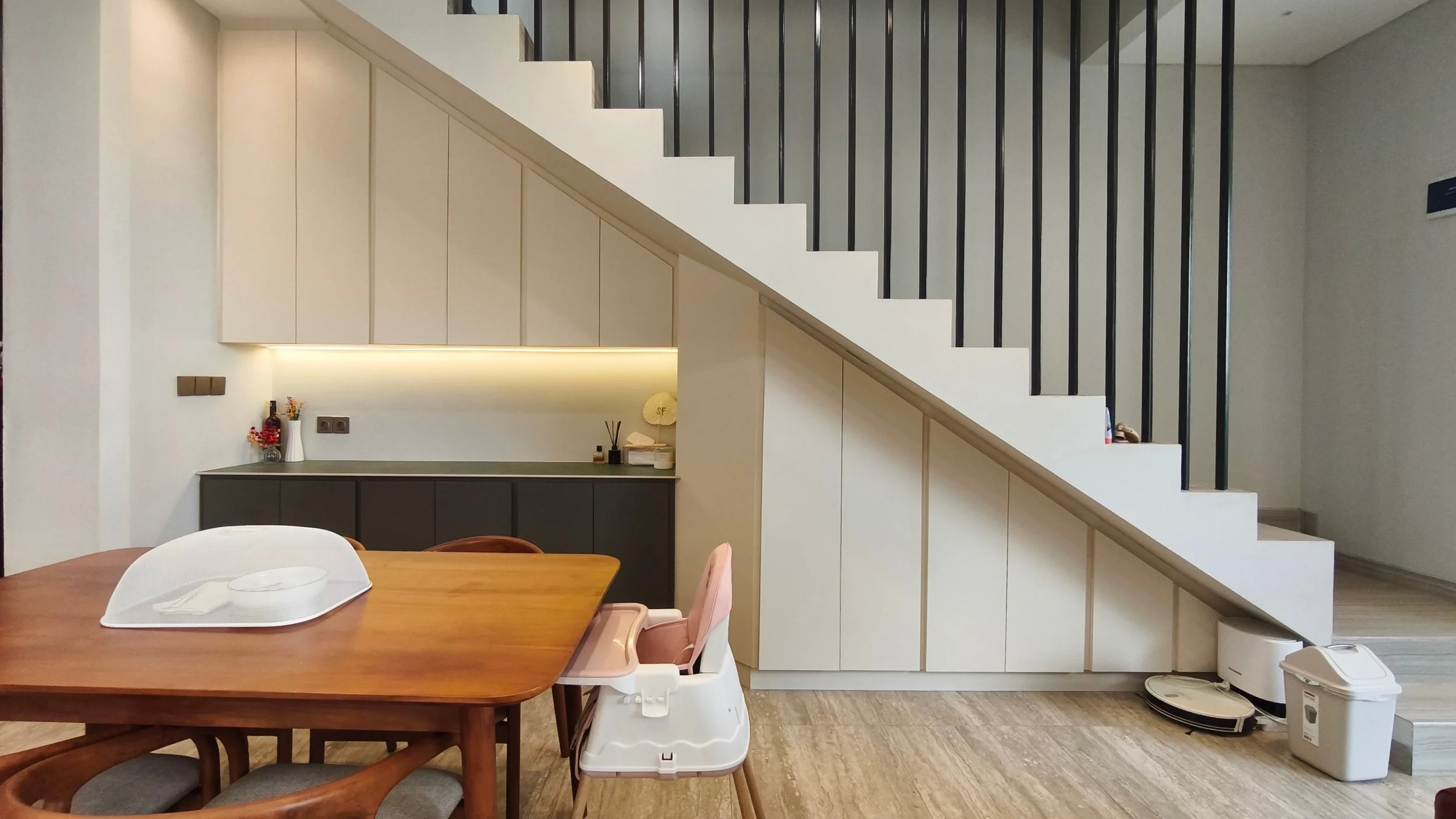 Interior view of a modern kitchen and dining area with a staircase. The wall features light-colored cabinets with hidden handles, a countertop with decorative items, including vases and a lamp, and a black lower cabinet. A wooden dining table with wh