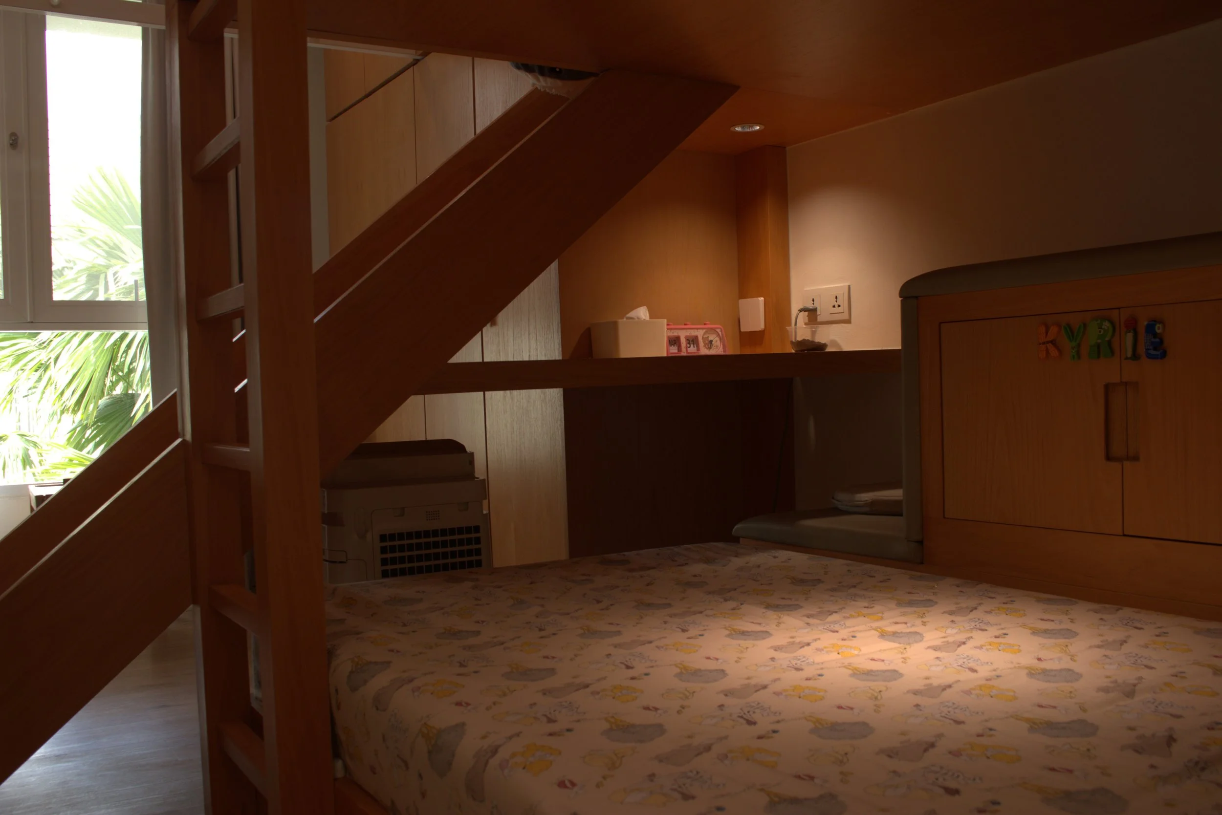 Interior of a small room with wooden stairs, a window with blinds showing green foliage outside, a bed with a patterned cover, a desk with items including tissue box and a calendar, and a cabinet with colorful magnetic letters that spell 'KYLE'.