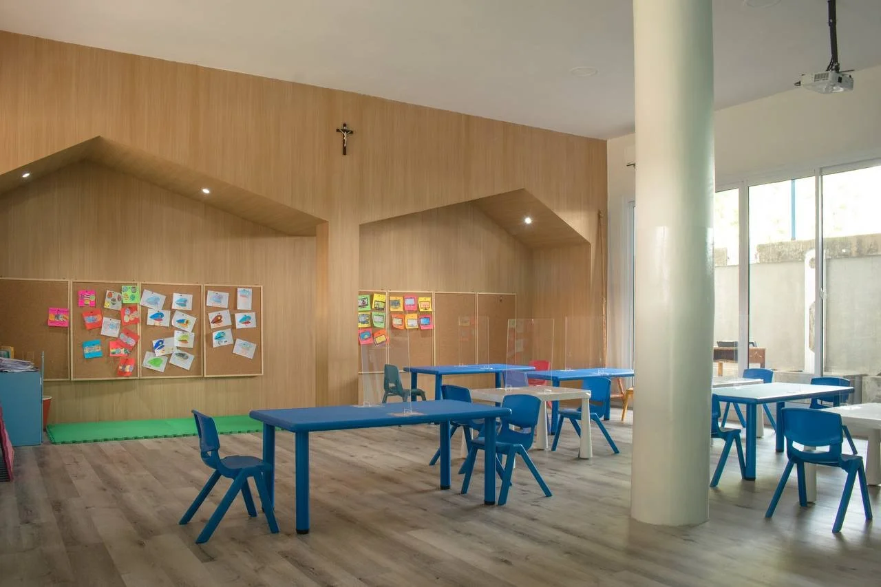Empty classroom with blue desks and chairs, a large white pillar, and a wooden wall decorated with colorful children's artwork, near a window with natural light.