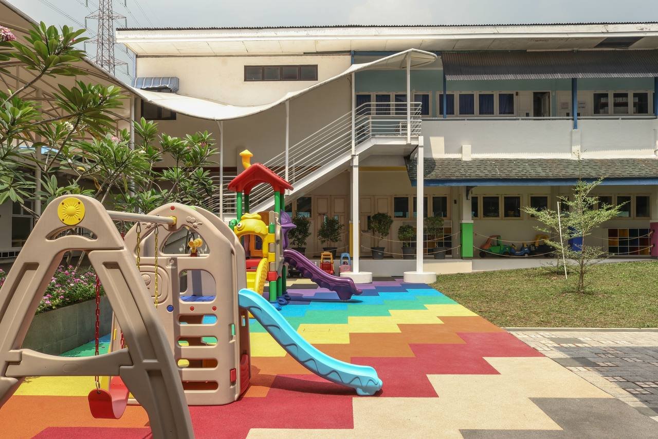 Colorful playground with slides and swings in front of a building with a rooftop staircase and a rainbow-colored mat on the ground.