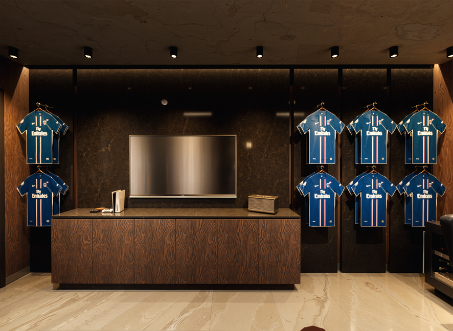 Display of six blue and white Real Madrid jerseys hanging on a black wall behind a wooden reception counter, with a television screen in the center.
