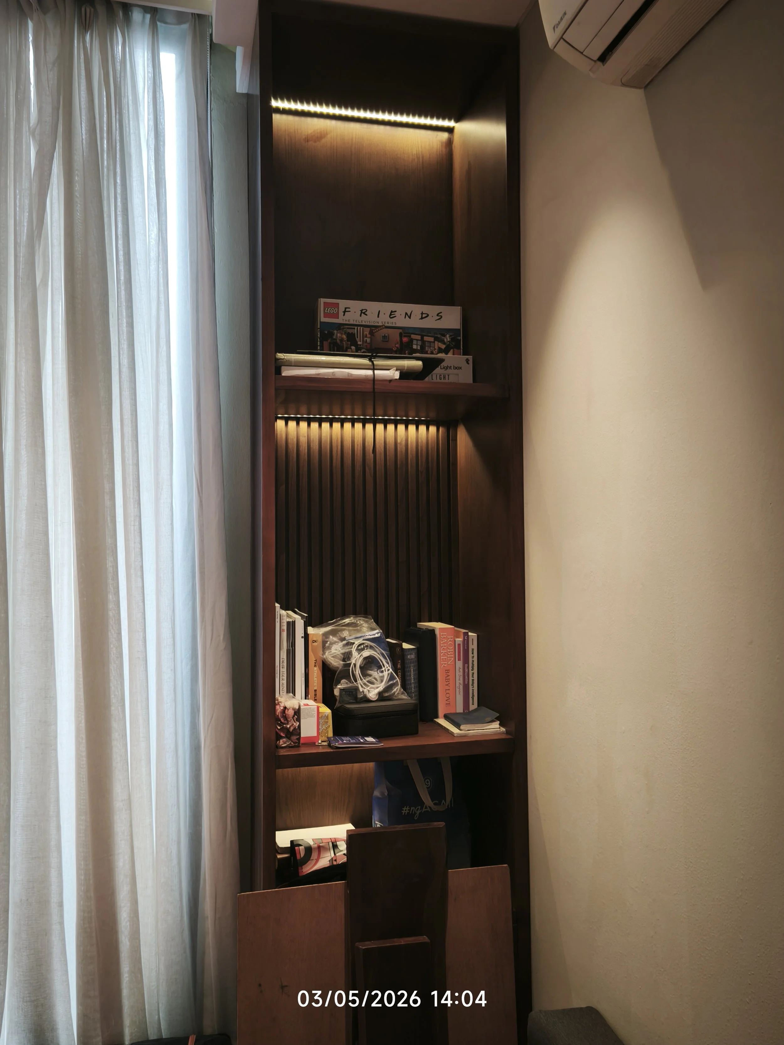 A tall wooden bookshelf beside a window with sheer white curtains, filled with books and various objects.