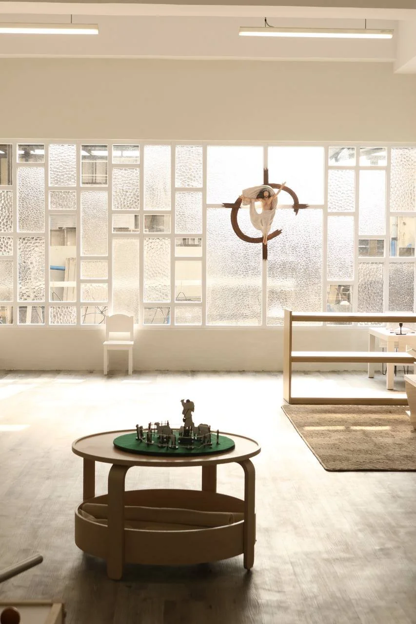 Bright, minimalist room with a wooden table displaying a small model puzzle, a white chair, large frosted windows, and a woman in a white dress hanging from a circular wooden orb suspended from the ceiling.