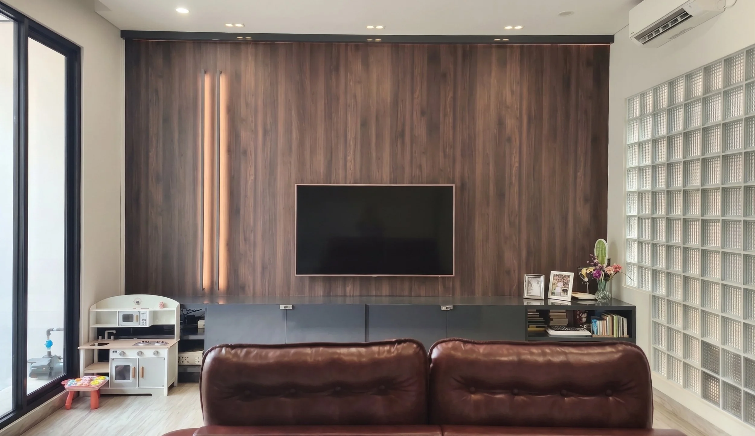 Living room with a wood accent wall, mounted flat-screen TV, glass block wall on the right, a side table with framed photos and a flower vase, brown leather chairs, and children's play area with a white toy kitchen.