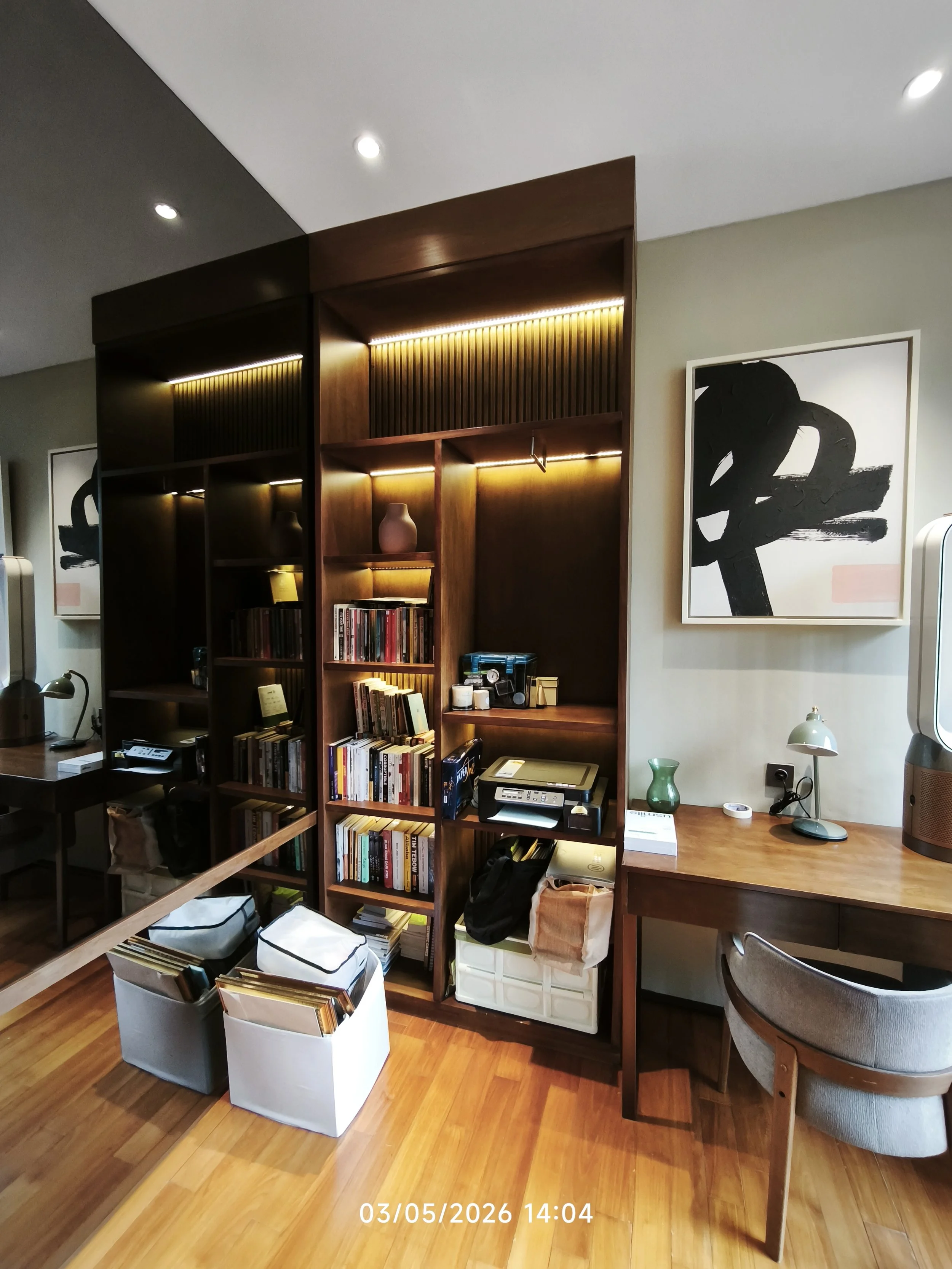 A corner of a room with a wooden bookshelf filled with books, a desk with a printer, a lamp, and various items, framed artwork on the wall, and two white storage boxes and a magazine holder on the floor.