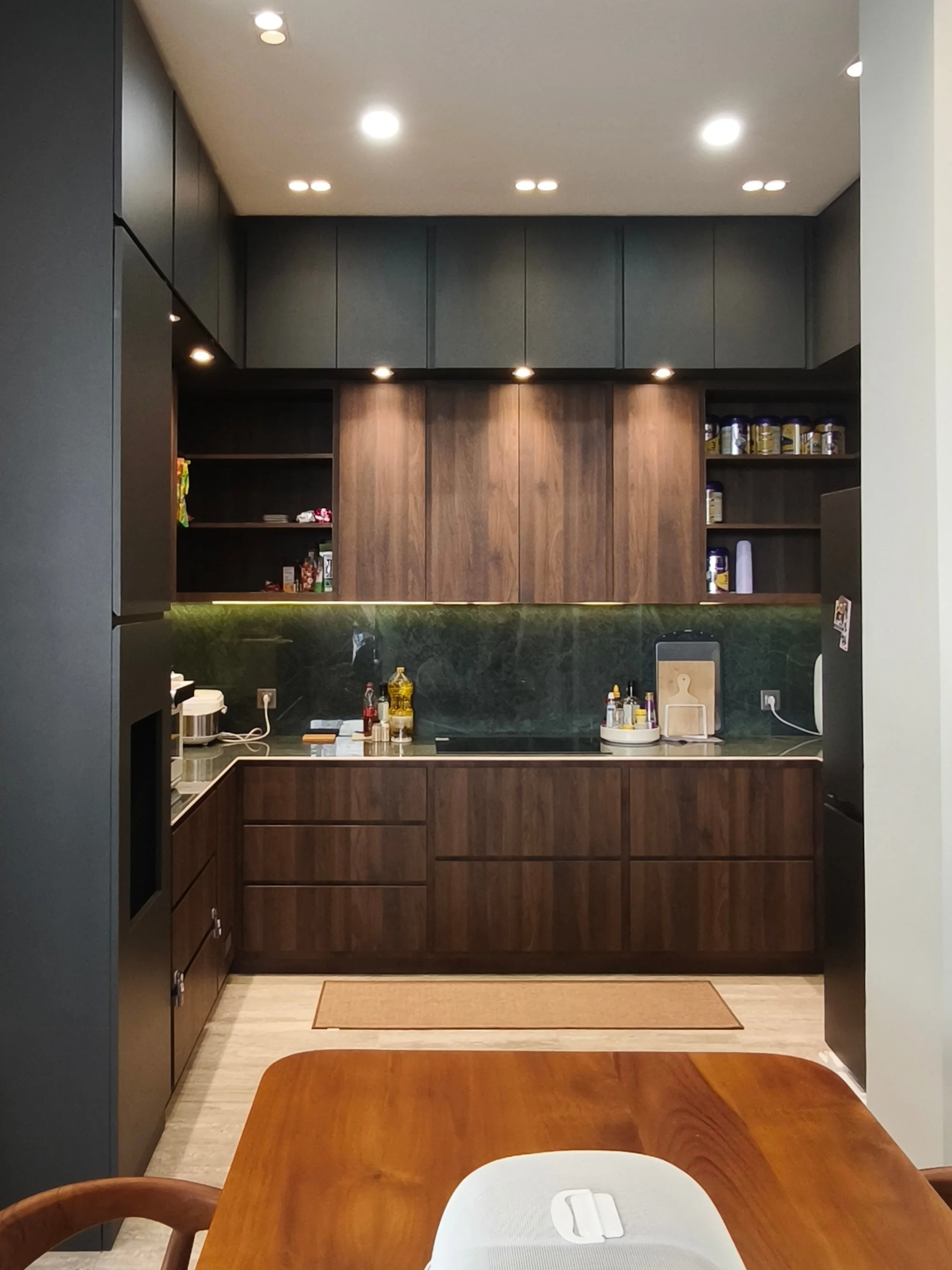 View of a modern kitchen with dark upper cabinets, wooden lower cabinets, a black countertop with a green backsplash, and various kitchen items on the counter. A wooden dining table with a chair in the foreground and a white dish drying rack on the t