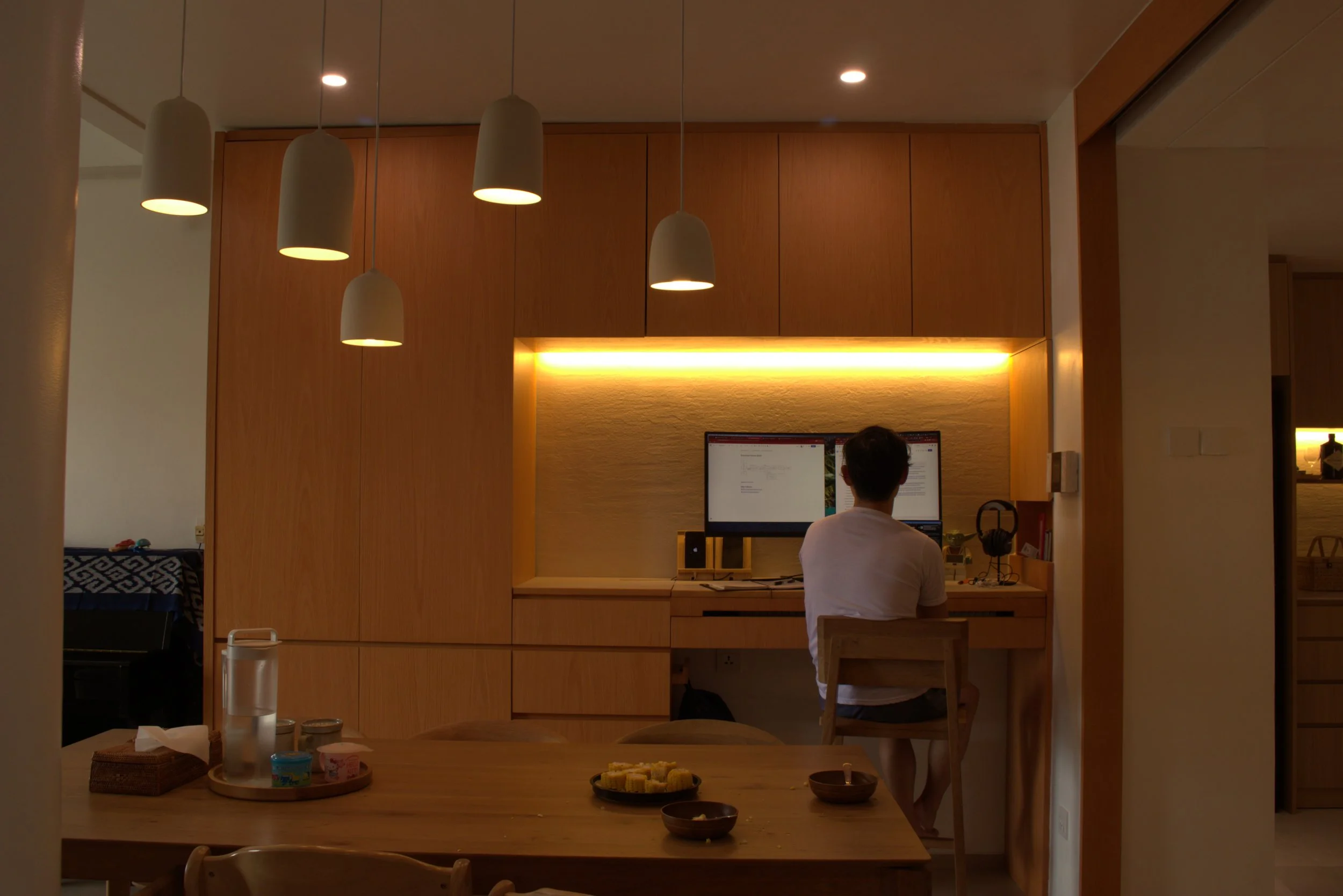 A person sitting at a desk in a modern home office with wooden cabinetry, illuminated by hanging pendant lights and recessed ceiling lights, working on a dual-monitor computer setup.
