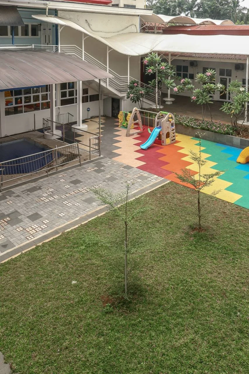 Outdoor playground area with a slide, climbing structure, and colorful rubber flooring, surrounded by trees and a grassy lawn, adjacent to a building with covered walkways.