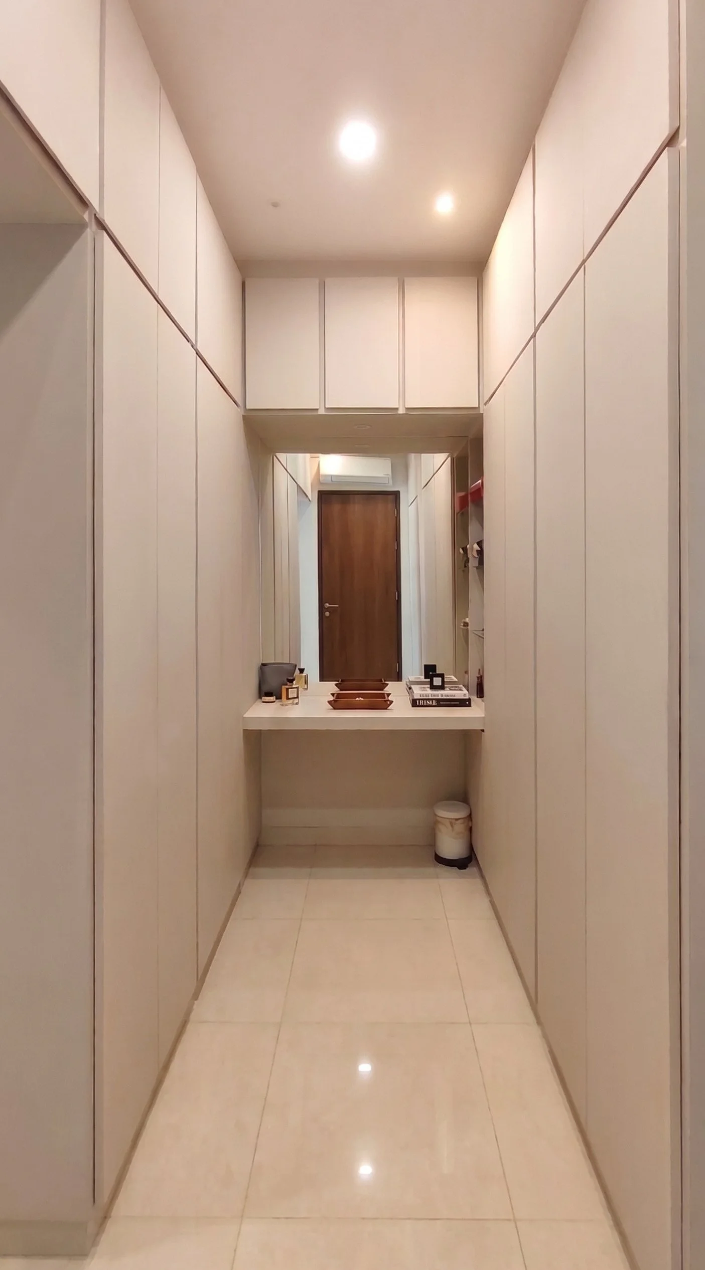 A narrow hallway with white walls, beige tiled floor, and a wooden door at the end. There is a small white counter with decorative items, books, and a mirror above it.