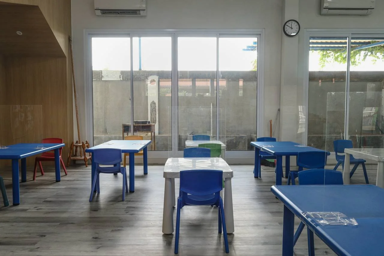 Empty classroom with blue and green chairs and tables, large windows with outdoor view, wall clock, and air conditioning units