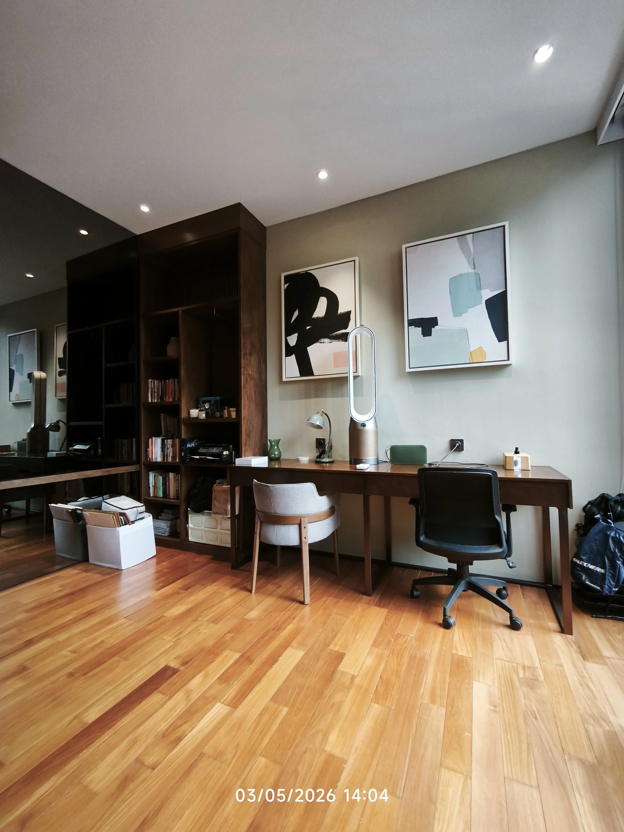 A home office setup with a wooden desk, a black office chair, and a white chair. There is a Dyson air purifier, a small lamp, books, and various office supplies on the desk. On the wall, there are two framed abstract artworks. The room has wooden flooring and a bookshelf with books and decor. The ceiling has recessed lighting.