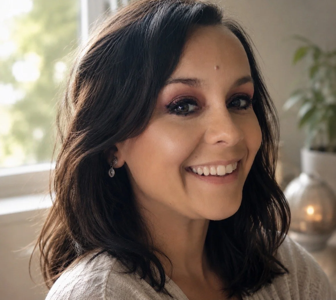 Femme souriante avec maquillage et boucles d'oreilles, arrière-plan de fenêtre et plante.