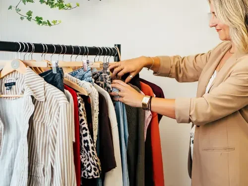 A woman standing in front of a clothing rack with pink and tan clothes, smiling in a boutique or store.