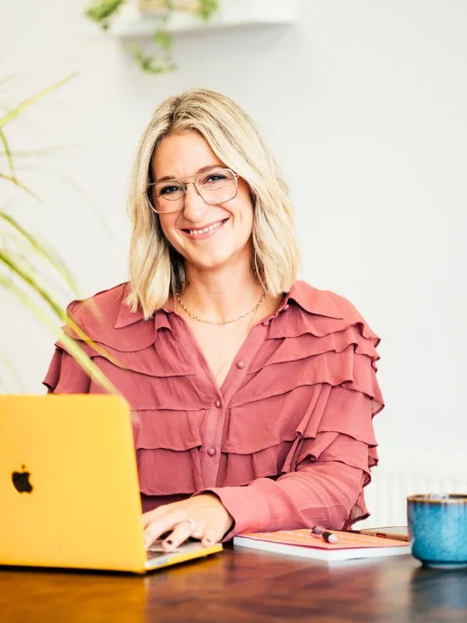 A woman with blonde hair and glasses smiling at a yellow laptop, sitting at a wooden desk with a notebook and a mug.