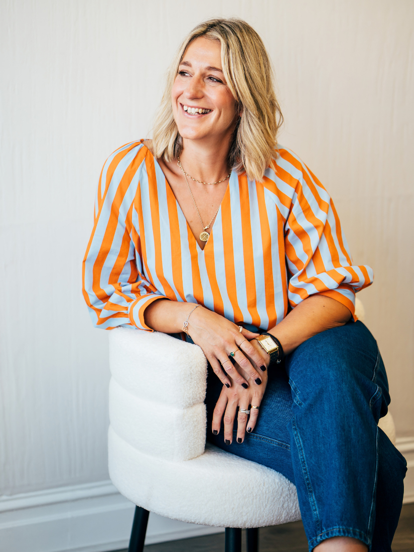A woman with blonde hair smiling, wearing an orange and white striped shirt, sitting on a white textured chair.