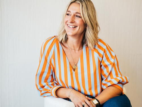 A smiling woman with blonde hair wearing a striped orange and white blouse sitting against a light-colored background.