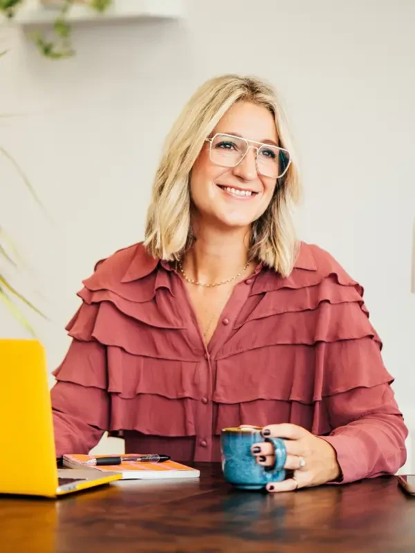 A woman with blonde hair and glasses sitting at a desk, smiling, with a yellow laptop and a blue mug in front of her.