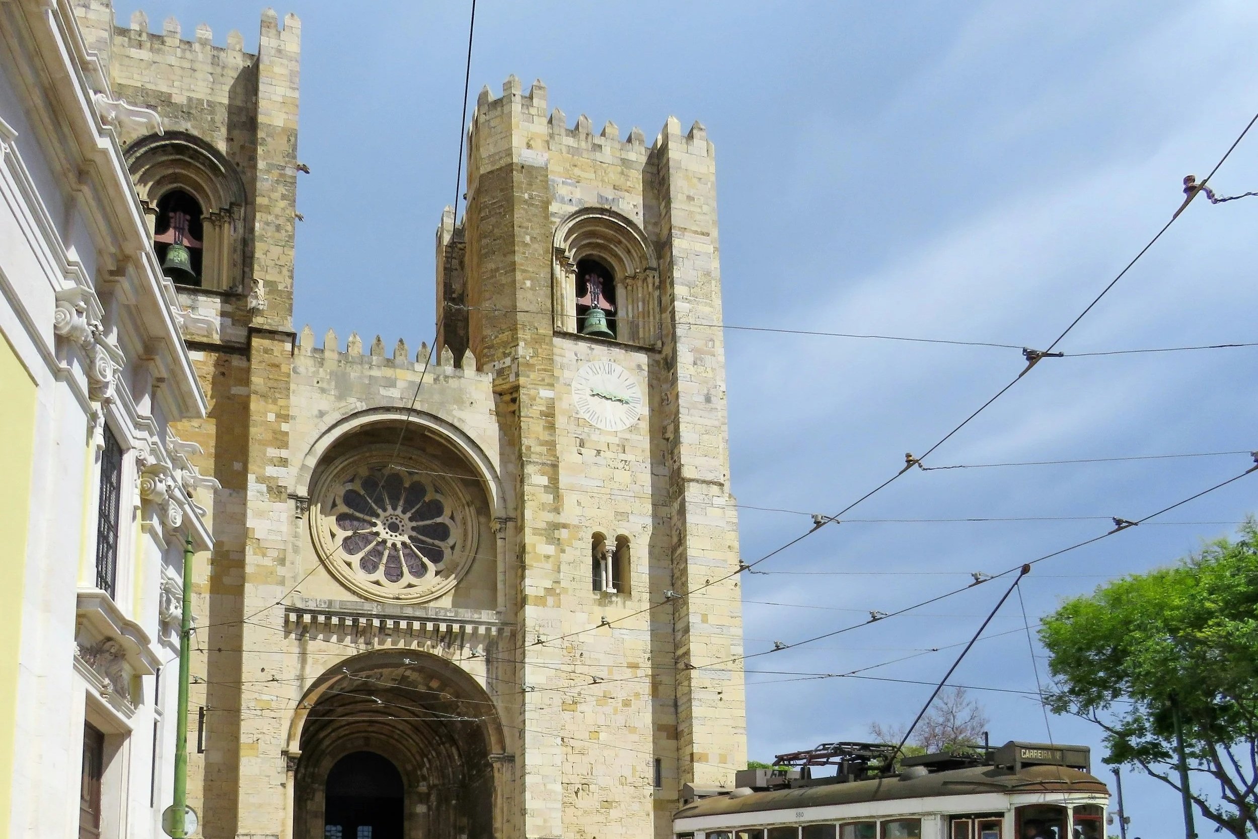 A historic church with a tall stone clock tower and bell, set against a blue sky. Tram wires cross the scene, and part of a tram is visible at the bottom.
