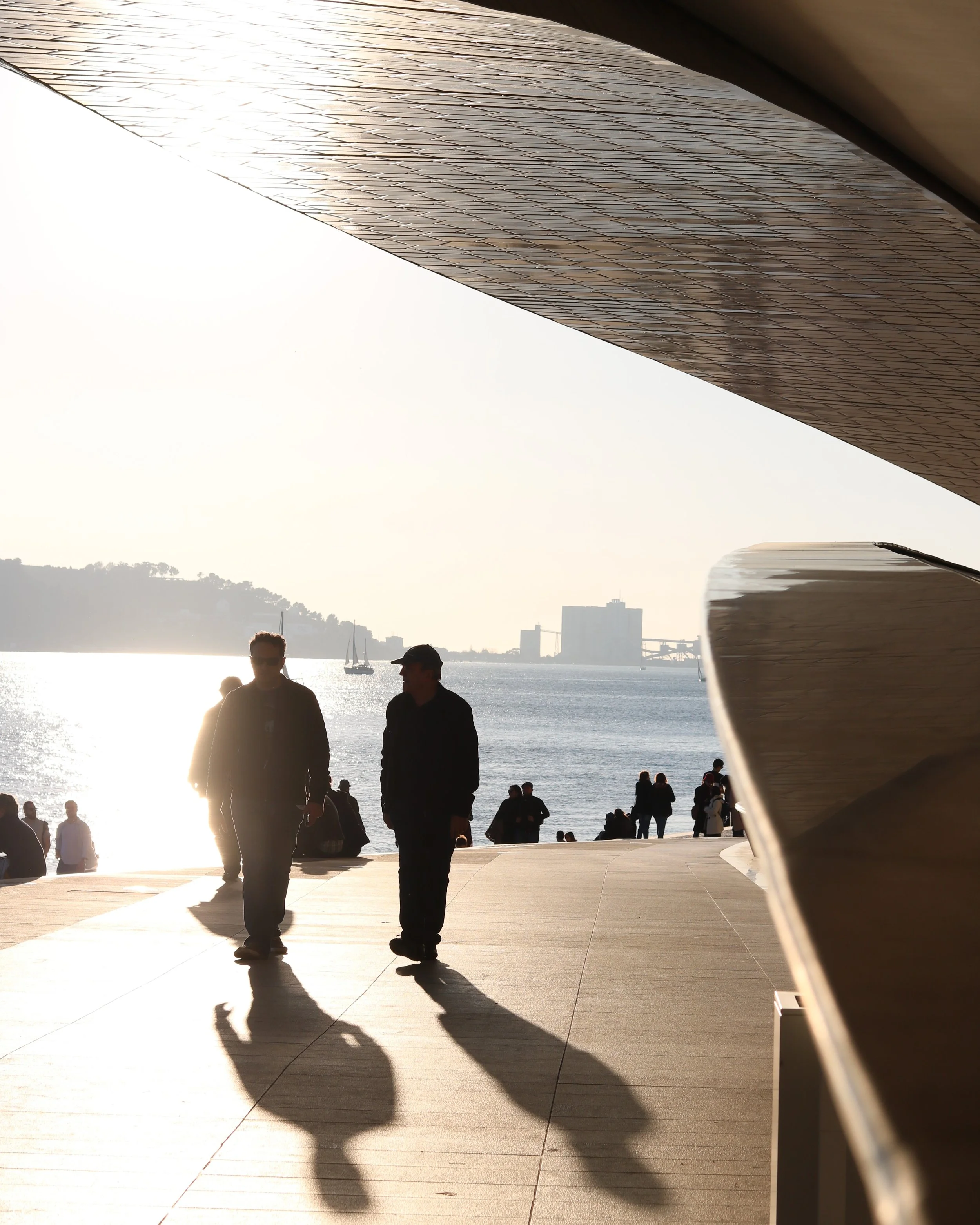 People walking on a waterfront promenade with the sun setting against a city skyline and boats sailing in the water.