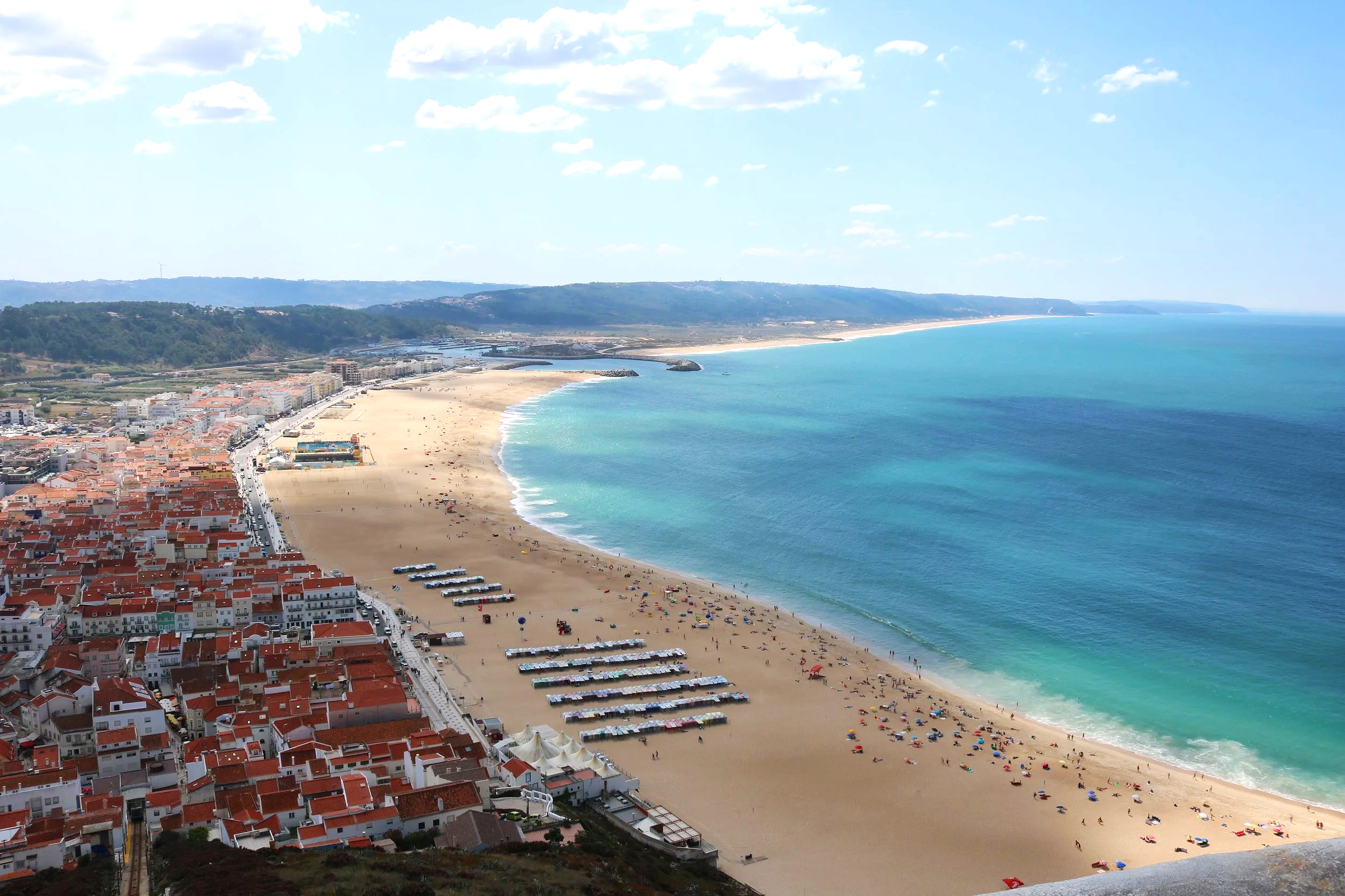 Coastal town with red-roofed houses, a sandy beach with colorful umbrellas, and the ocean under a partly cloudy sky