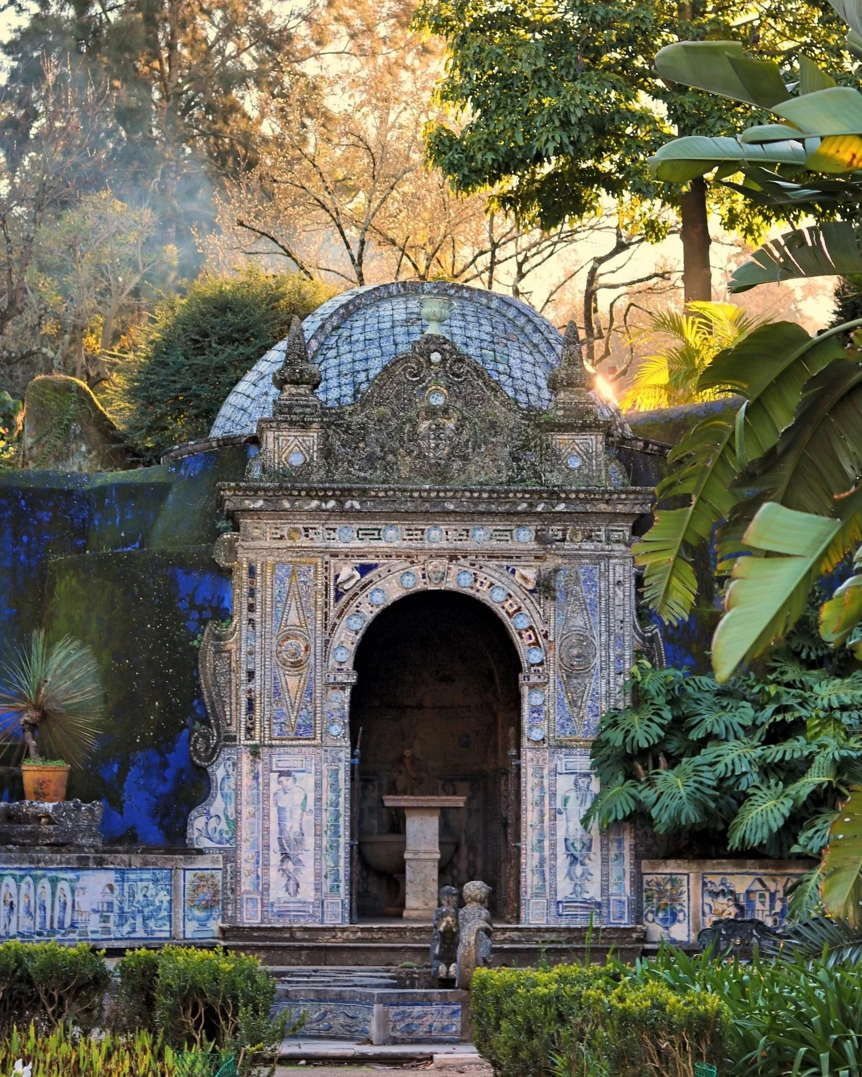 A decorative mosaic archway in a garden, adorned with colorful tiles and surrounded by lush greenery, with sunlight filtering through trees in the background.