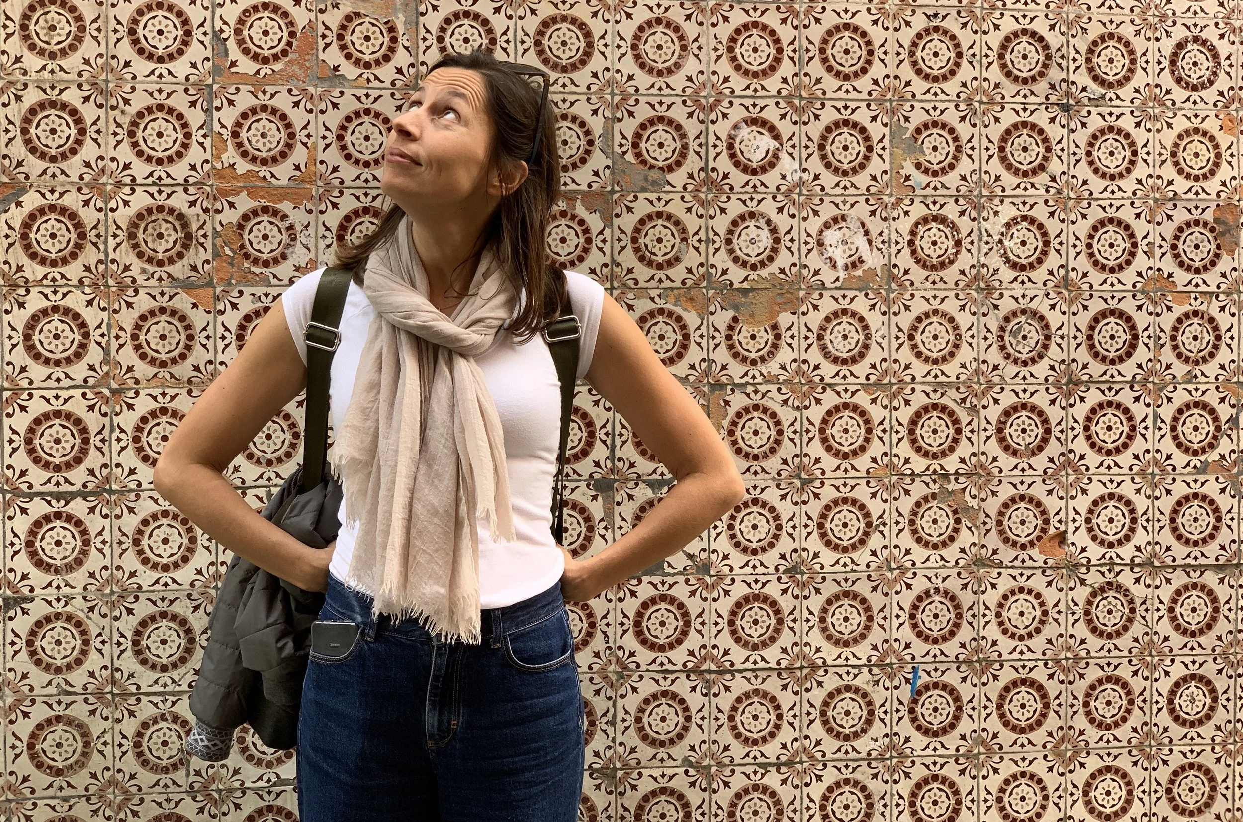Woman standing in front of a decorative tiled wall, looking upward with her hands on her hips, wearing a white t-shirt, a beige scarf, and carrying a backpack.